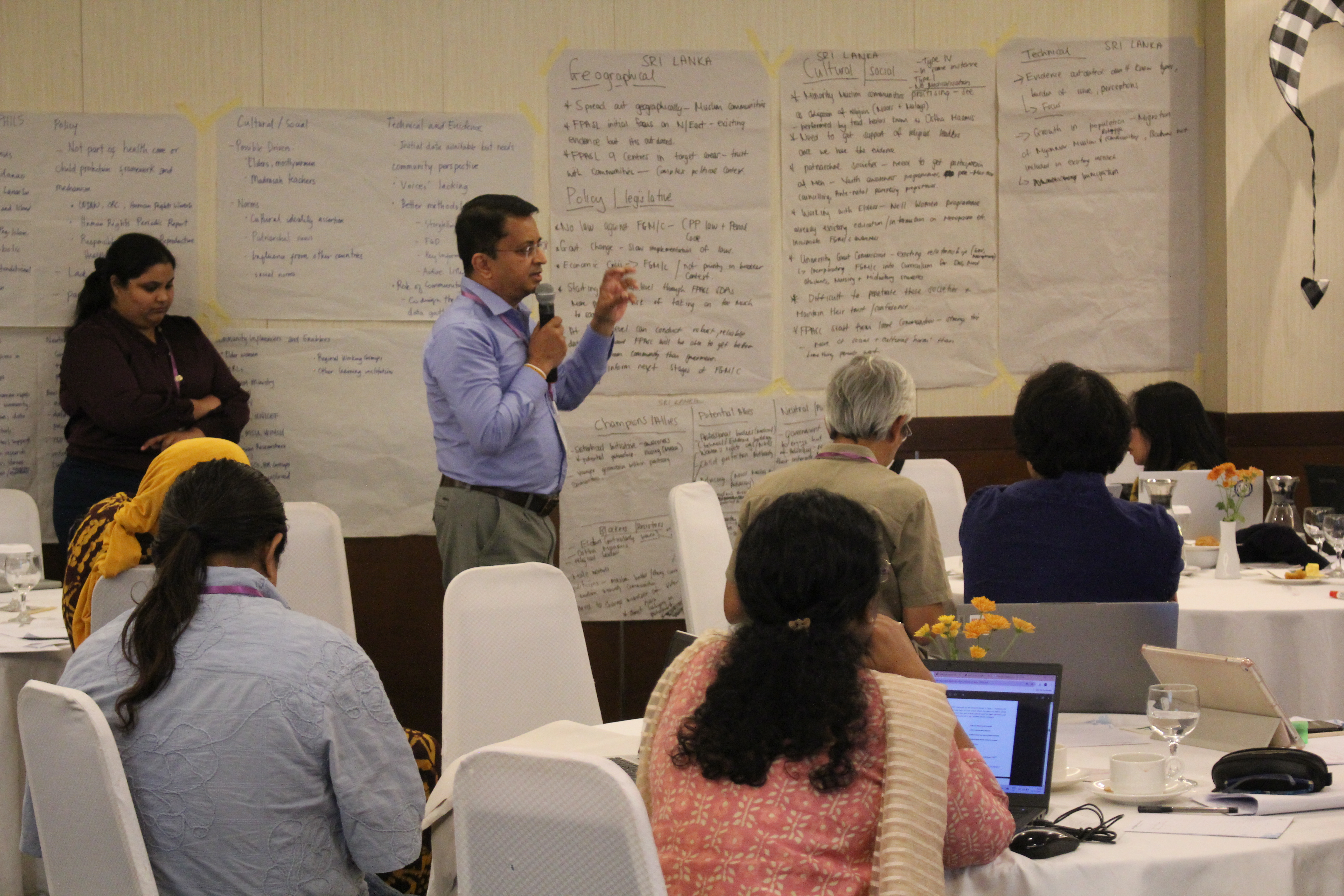 A man holding a microphone speaks to a small group seated at round tables in a conference room, while large sheets of handwritten notes are taped to the wall behind him. Participants listen and take notes on laptops, creating a collaborative workshop atmosphere.