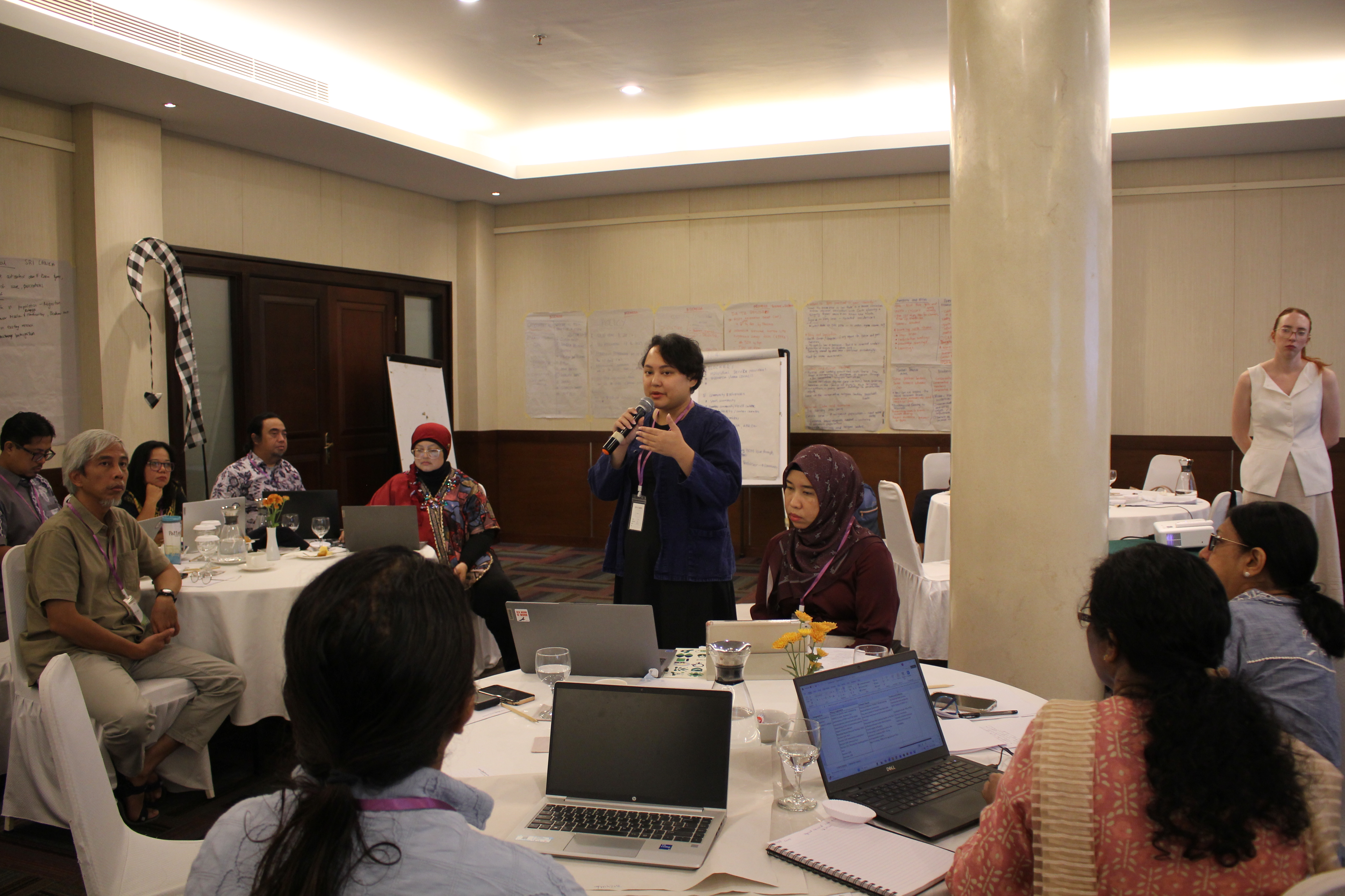 A woman stands and speaks into a microphone during a workshop session in a hotel conference room, while other participants sit at round tables with open laptops and notebooks. Handwritten charts are displayed on the walls, and attendees listen attentively.