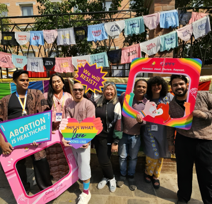 A diverse group of people poses outdoors holding colorful signs with messages like “Abortion is Healthcare,” “This is what love looks like,” and “We will resist.” Behind them, underwear with activist slogans hangs on a line. The scene is joyful and celebratory, promoting LGBTQ+ and reproductive rights, with IPPF and ILGA branding visible.