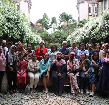 A diverse group of around twenty adults pose together in a garden courtyard surrounded by lush greenery and flowering vines. Some are seated on a low stone ledge while others stand behind them, smiling and gesturing toward the camera in a relaxed, celebratory group photo.