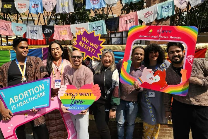 A diverse group of people poses outdoors holding colorful signs with messages like “Abortion is Healthcare,” “This is what love looks like,” and “We will resist.” Behind them, underwear with activist slogans hangs on a line. The scene is joyful and celebratory, promoting LGBTQ+ and reproductive rights, with IPPF and ILGA branding visible.
