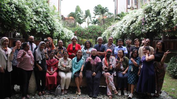 A diverse group of around twenty adults pose together in a garden courtyard surrounded by lush greenery and flowering vines. Some are seated on a low stone ledge while others stand behind them, smiling and gesturing toward the camera in a relaxed, celebratory group photo.